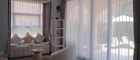 A modern living room with a sofa, a round coffee table, and a white armchair. There is a large window with sheer curtains and a view of outdoor furniture.