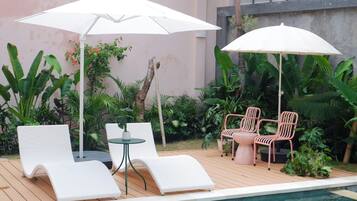 A poolside area with white lounge chairs, a small round table, and two white umbrellas providing shade.