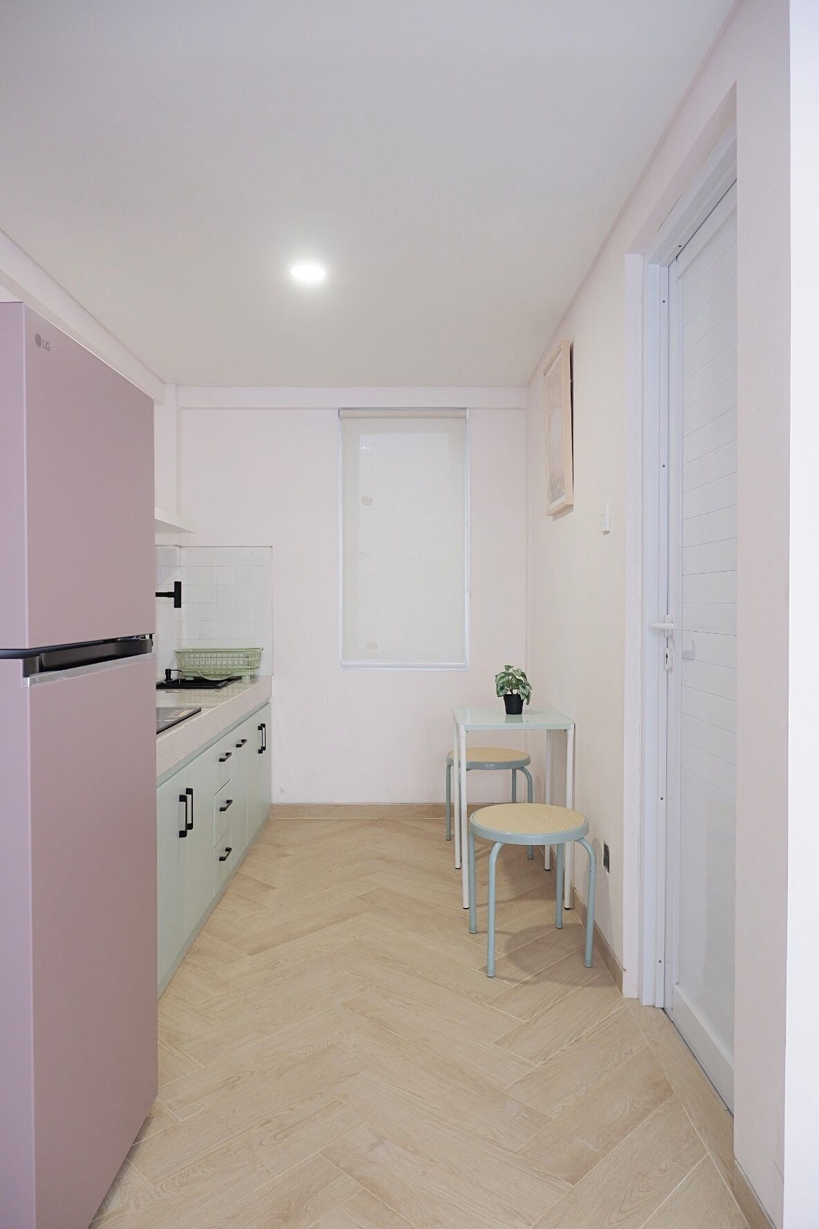 A compact kitchen with a refrigerator, a sink, and a window. There is a small table with a chair and a potted plant.