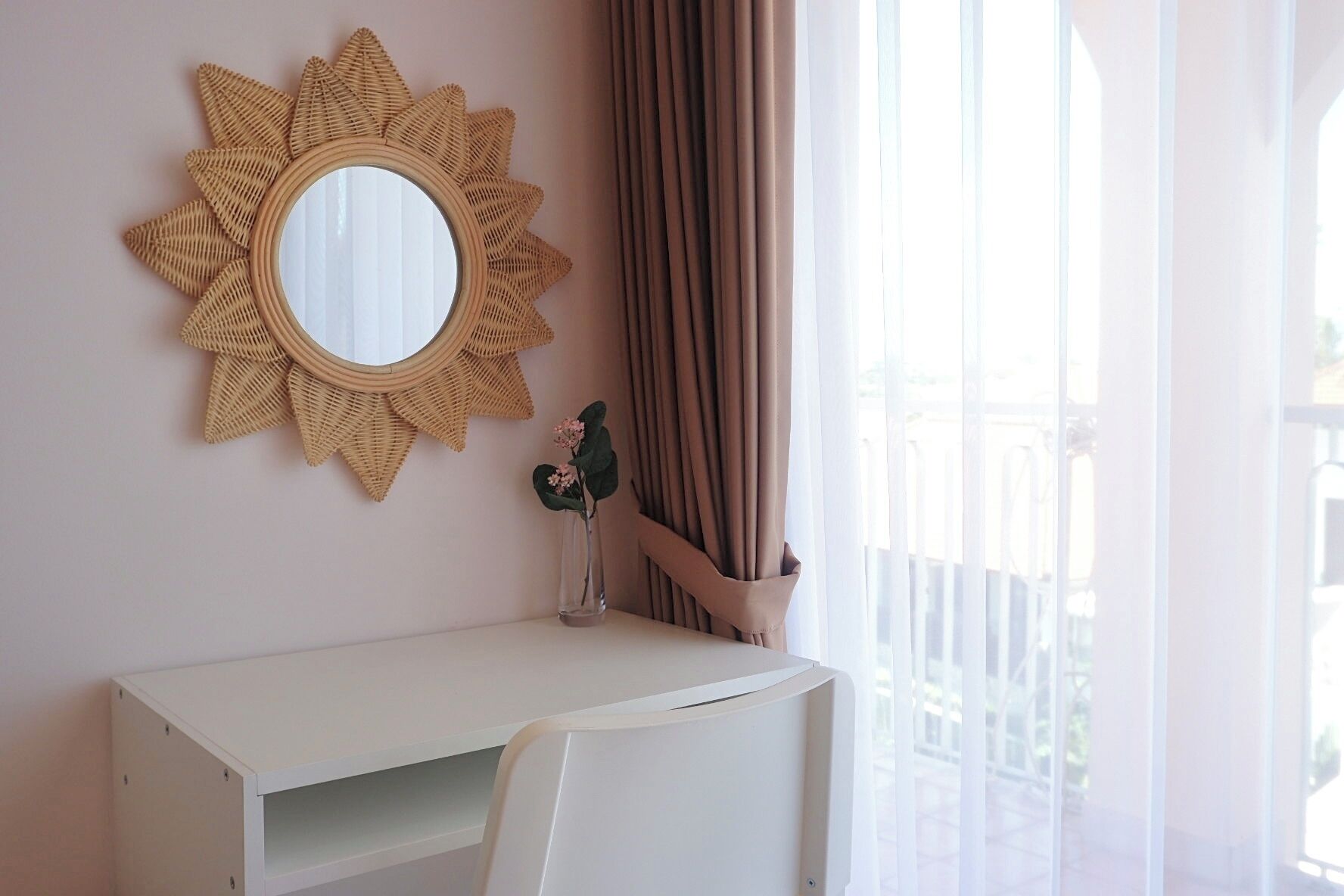 A room with a white desk, a round mirror with a sunburst frame, a vase with flowers, and a window with curtains.