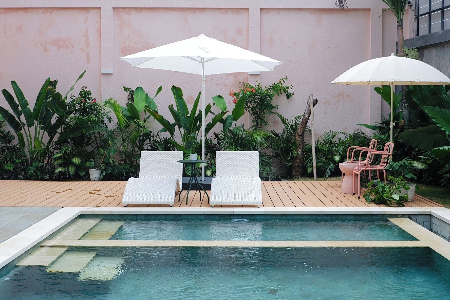 A pool area with two white lounge chairs and a table under white umbrellas, surrounded by lush tropical plants.