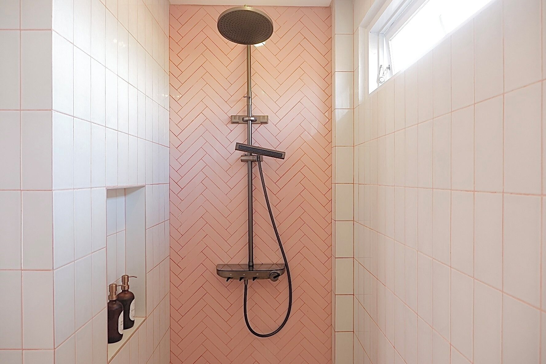 Une petite douche avec un mur orné d’un motif en chevrons, des murs recouverts de carreaux blancs et une étagère en verre fermée, où sont rangés les articles de toilette.
