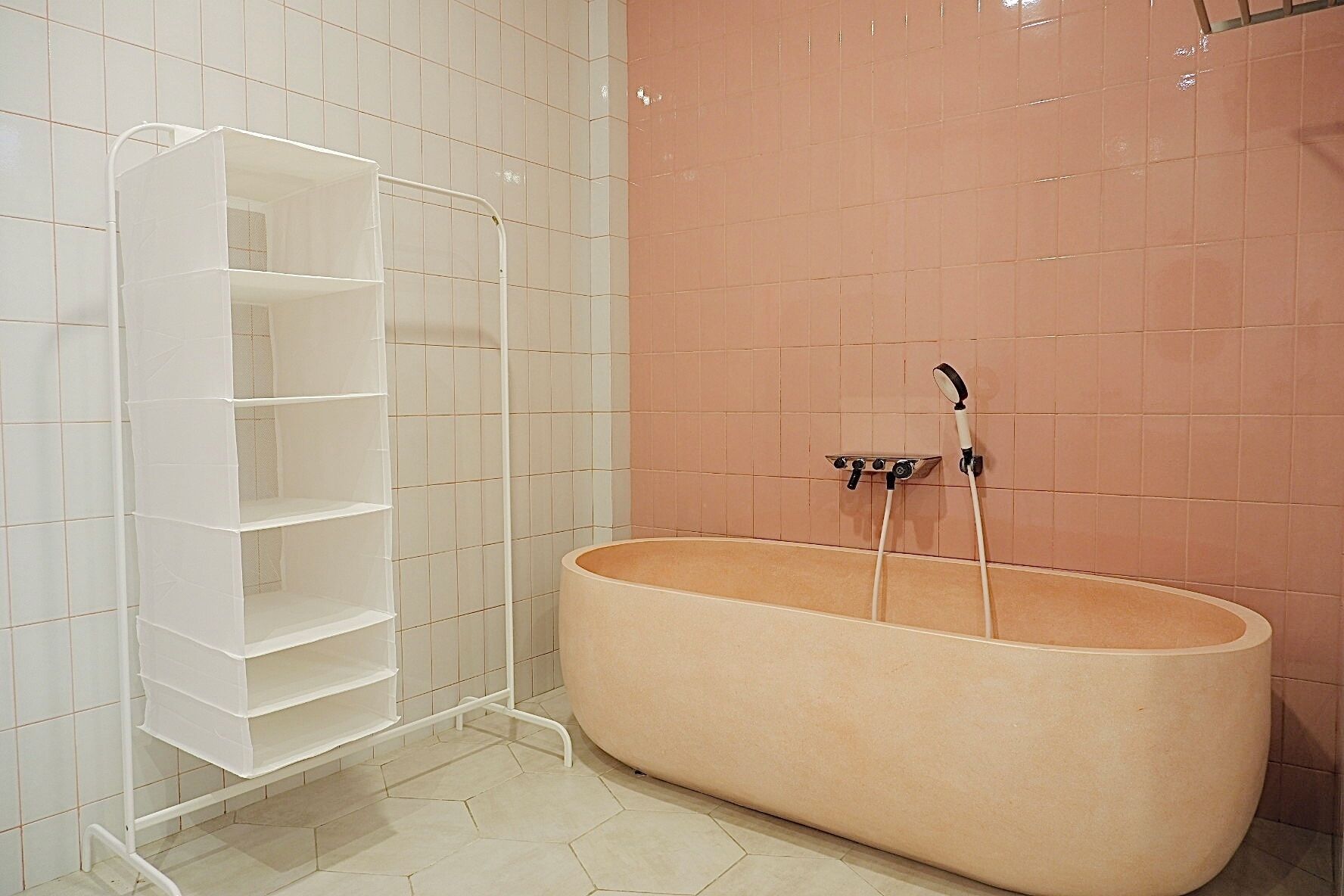 A bathroom with a large bathtub, a white shelving unit, and a pink tiled wall.