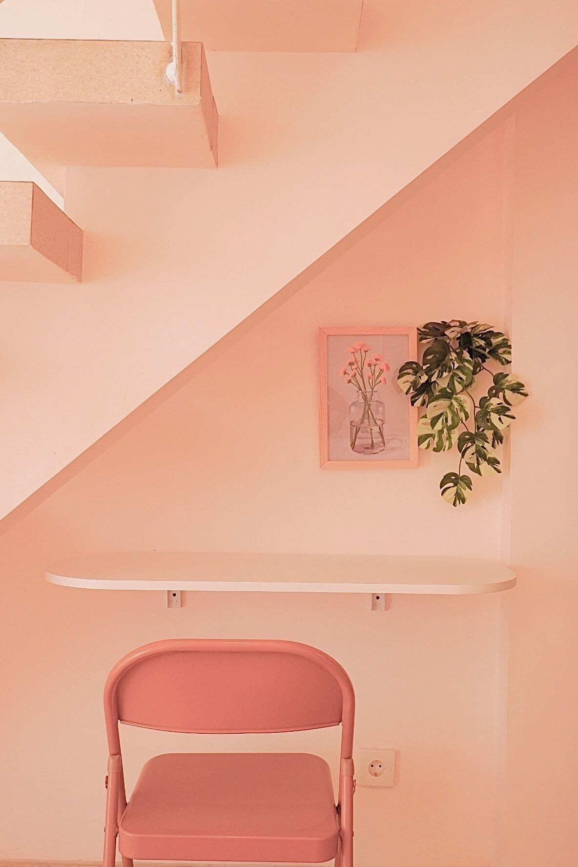 A pink chair in a room with a white shelf and a framed picture of flowers.