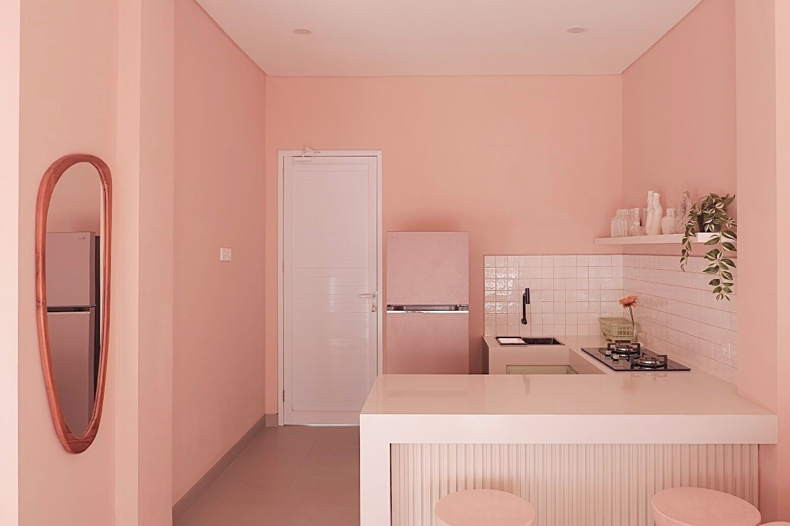 Eine kleine Küche mit weißer Arbeitsplatte, einem Spülbecken und einem Kühlschrank. An der linken Wand befindet sich ein Spiegel und auf der Arbeitsplatte ein Blumentopf.