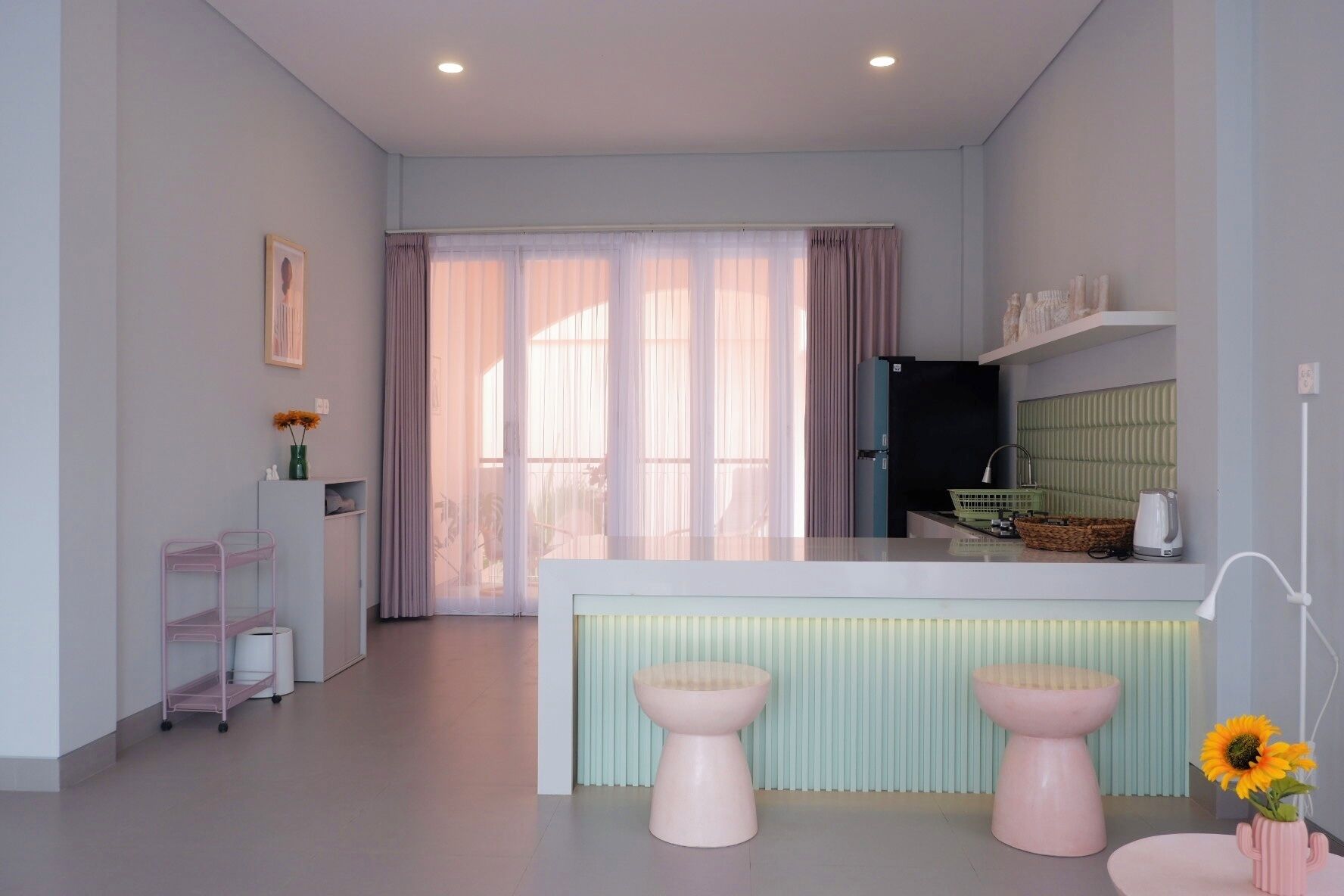 A modern kitchen with a central island, two pink stools, a sink, and a window with sheer curtains.