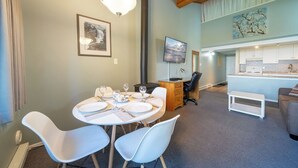 A modern room with a dining table set for four, a TV mounted on the wall, and a kitchen area visible through an open door.