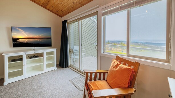 Une pièce avec un téléviseur posé sur un meuble blanc, un plafond en bois, une grande fenêtre donnant sur la mer et une chaise en bois avec un coussin orange.