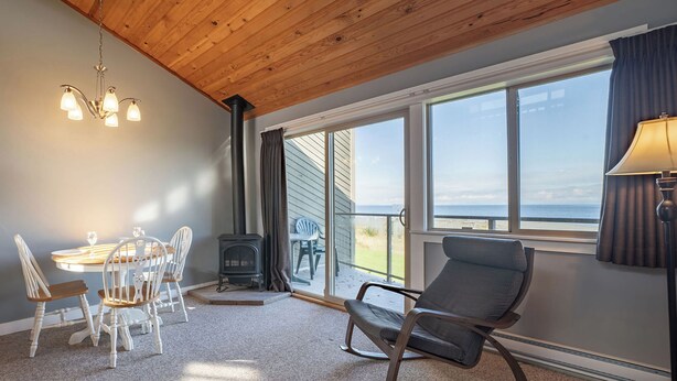 Une pièce chaleureuse avec un plafond en bois, une table avec des chaises, un fauteuil à bascule, une lampe et une grande fenêtre donnant sur la mer.