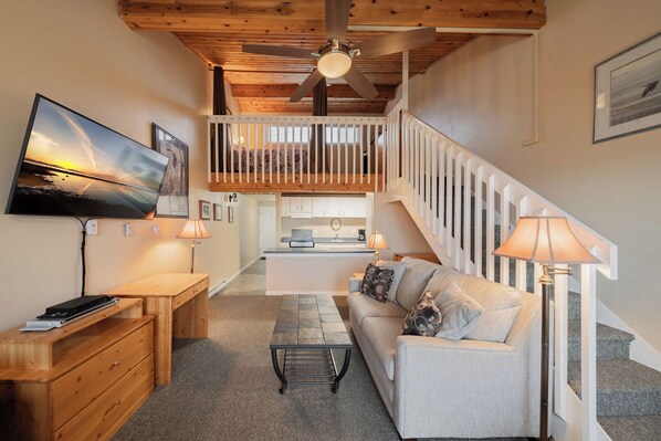 Una sala de estar acogedora con una televisión de pantalla plana, muebles de madera, una escalera y una cocina al fondo.