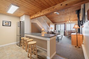 Una cocina con una isla central, taburetes de madera, un refrigerador y una escalera que lleva a un comedor con una mesa y sillas.