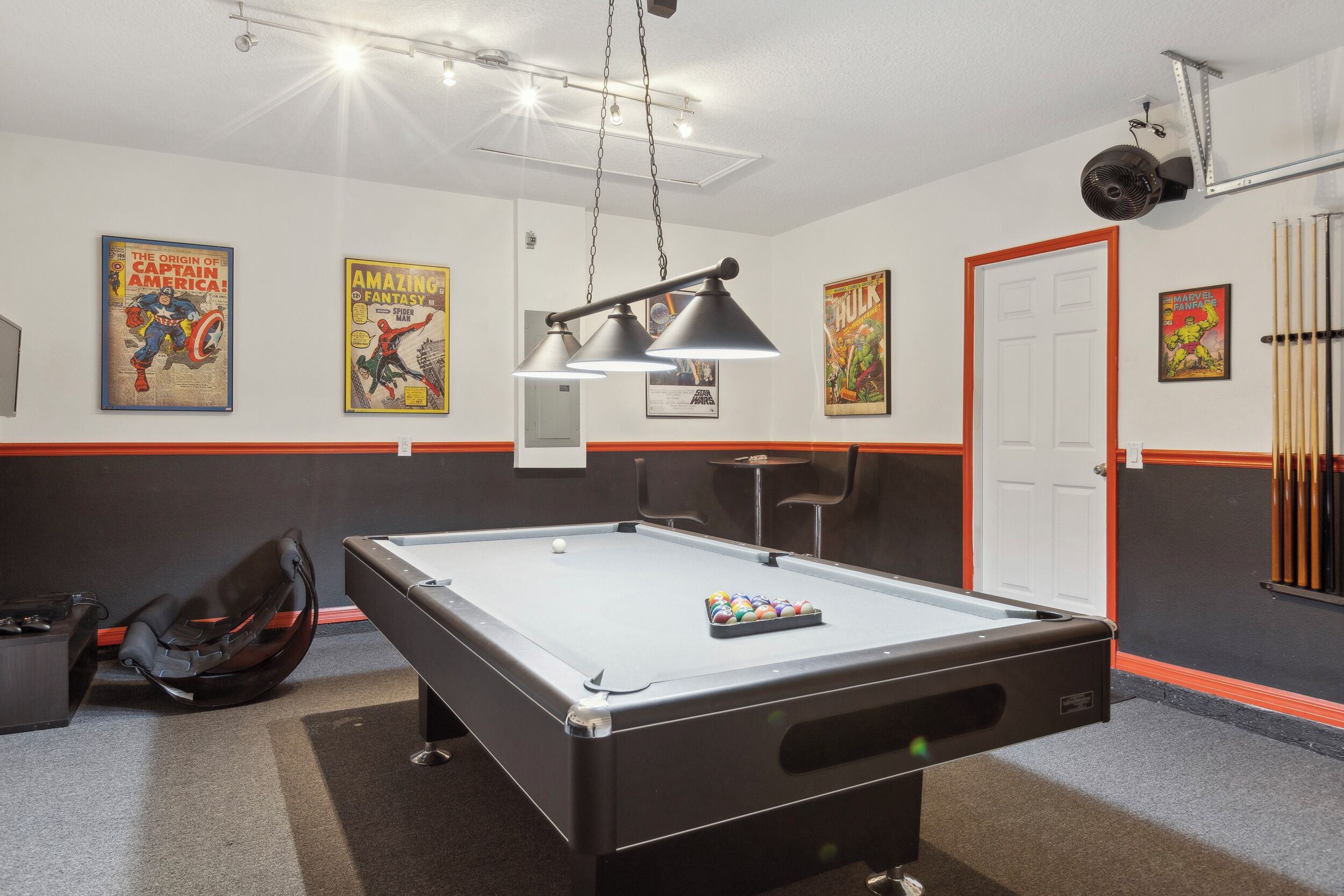 A pool table with a cue ball and a rack of billiard balls, a pool table chair, and a pool table bag. There are posters on the wall, a door, and a ceiling fan.