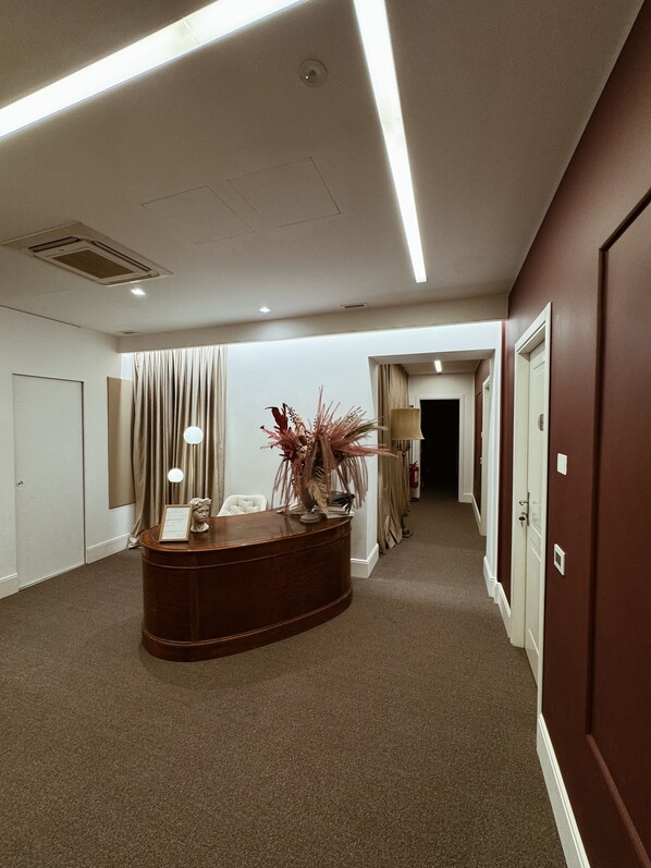 A hotel reception area with a wooden reception desk, a white chair, a vase with red flowers, and a lamp. - Residenza Imperiale - Luxury Guest House (Rome)