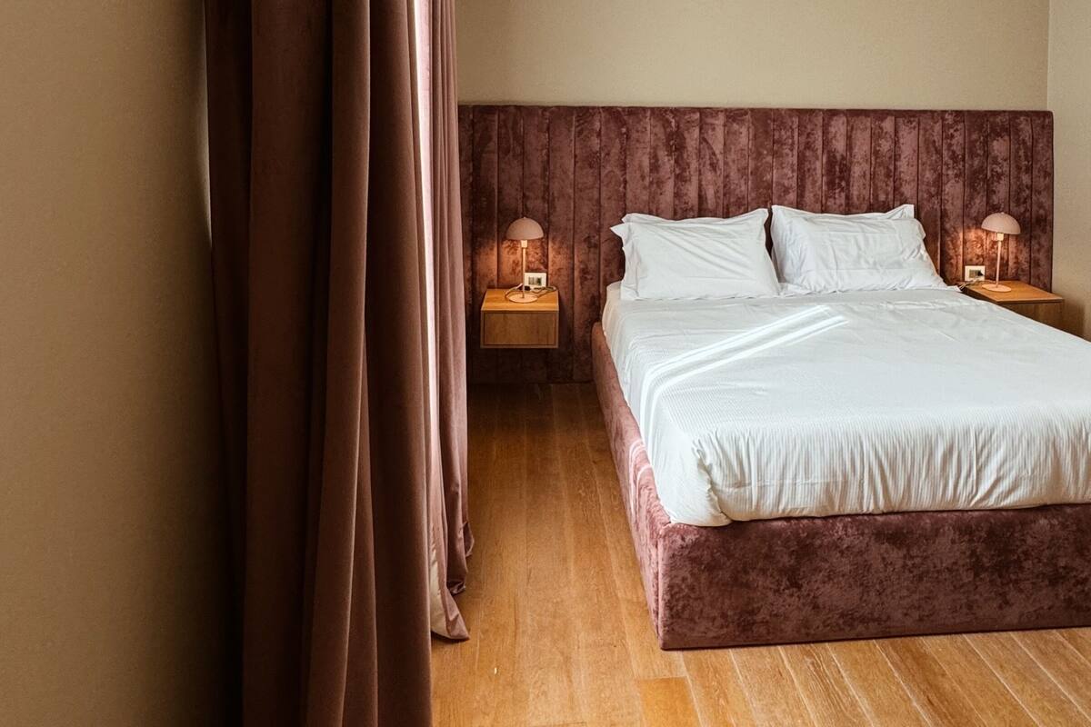 Un dormitorio con una cama grande, suelo de madera, cabecera, dos mesitas de noche con lámparas y una cortina.