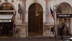 En bygning i klassisk arkitekturstil med en central indgang prydet med en detaljeret gavl og søjler. På hver side af indgangen findes butikker, herunder en bar og en butik med navnet 'TABACCHI'.
