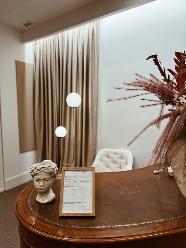 A room with a wooden table, a bust sculpture, a framed document, a white chair, and a large window with curtains.