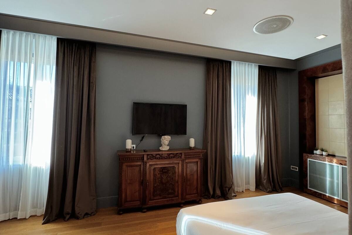 Habitación de hotel con una cama grande, suelo de madera, un televisor de pantalla plana sobre un mueble de madera y un ventanal con cortinas.