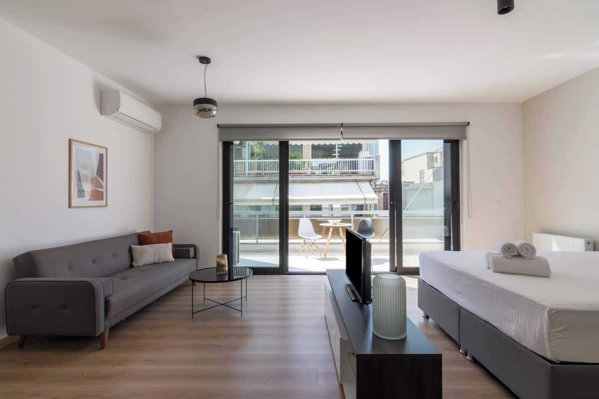 Een moderne woonkamer met een grijze bank, een klein rond tafeltje, een televisie en een bed met opgerolde handdoeken.