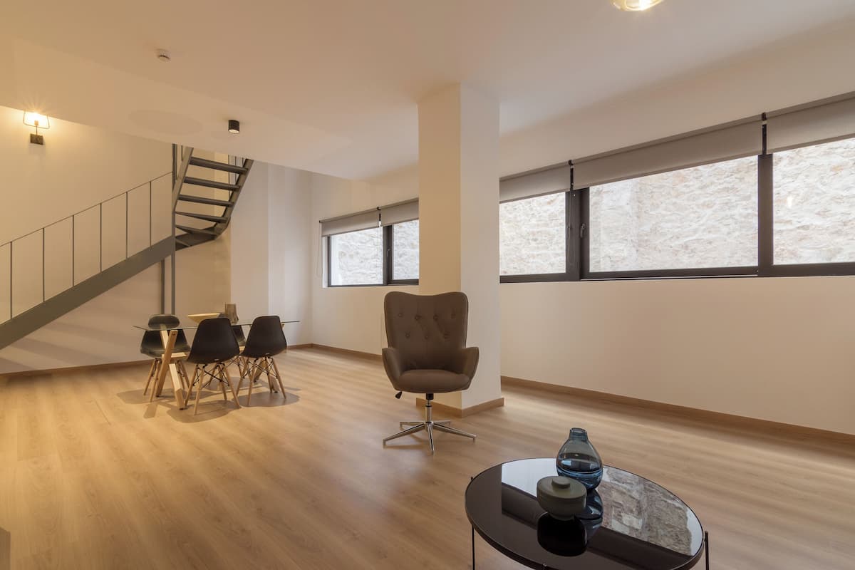 Ruime kamer met houten vloer, een eethoek met tafel en stoelen, een moderne fauteuil, en een trap.