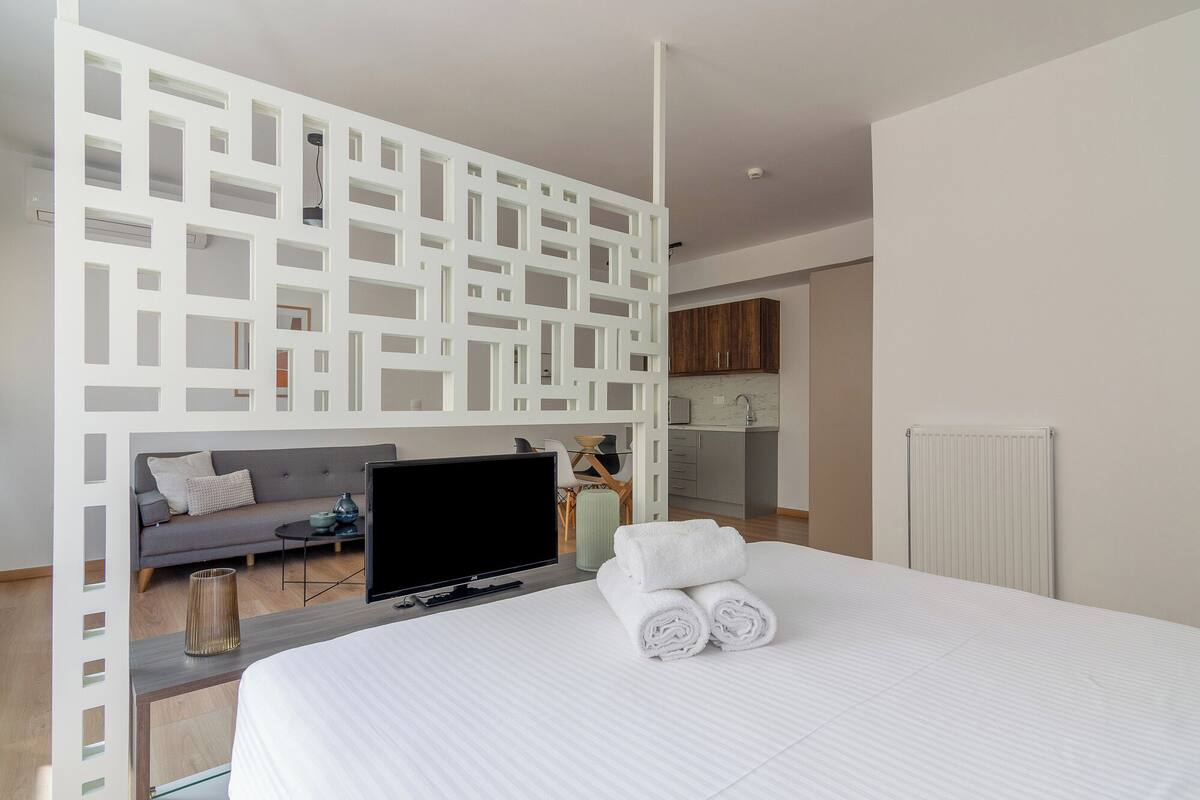 Een moderne hotelkamer met een groot bed, een televisie, een zithoek met een bank en een kleine tafel, en een keukenruimte zichtbaar door een open wand.