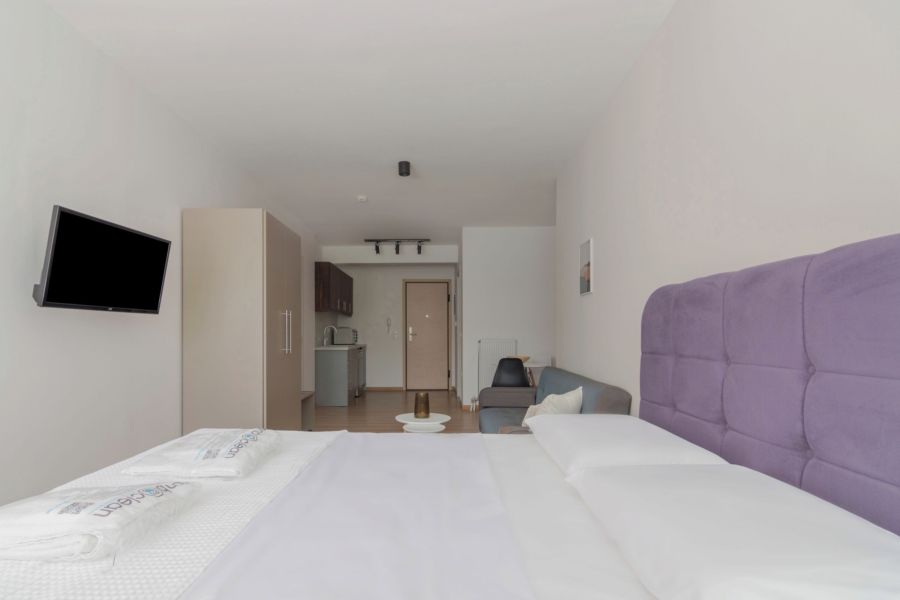 A hotel room with a bed, a television mounted on the wall, a kitchenette, and a bathroom visible through an open door.