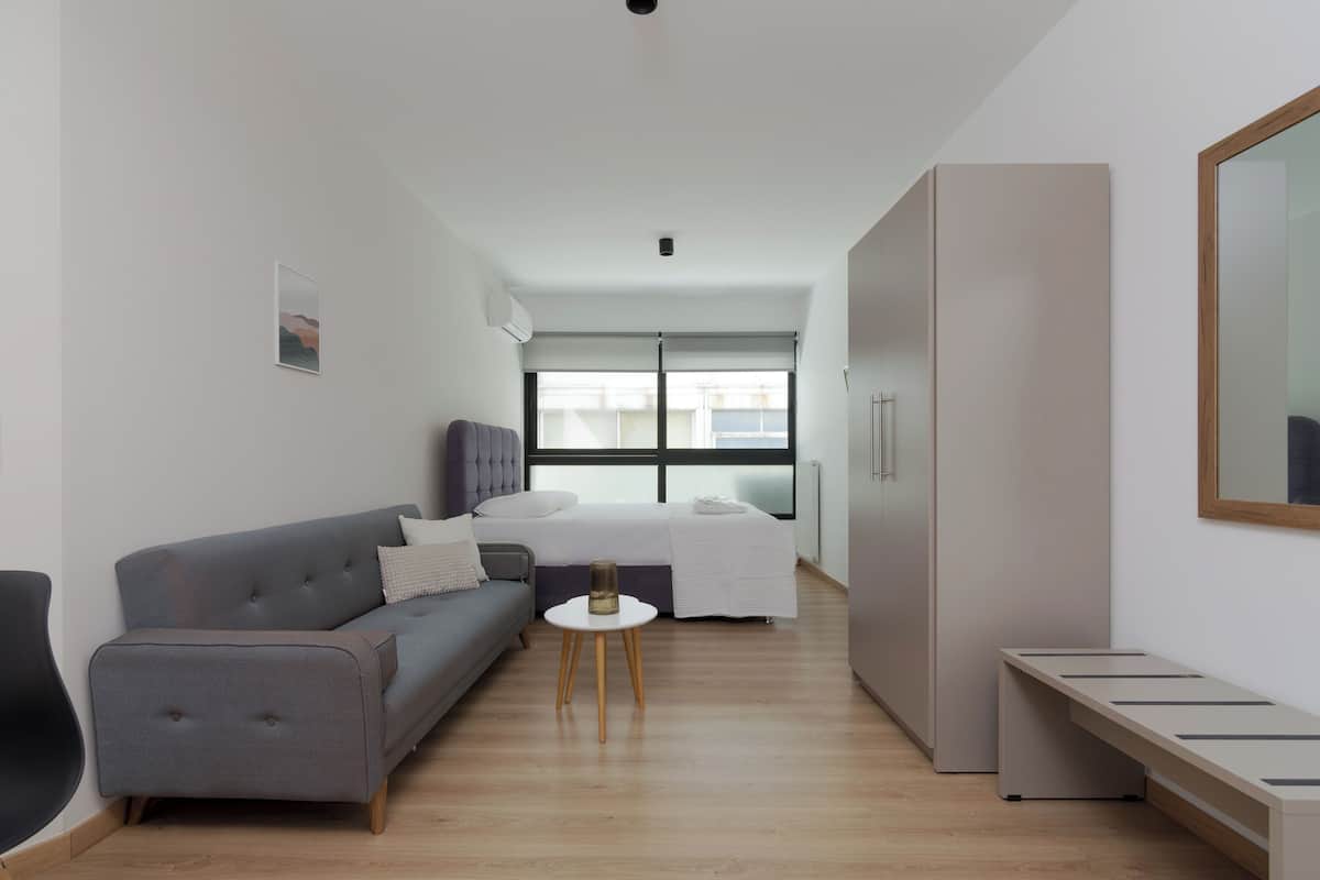 Een moderne, goed verlichte kamer met een grijze bank, een klein rond tafeltje, een bed met een witte dekbedovertrek en een kledingkast.