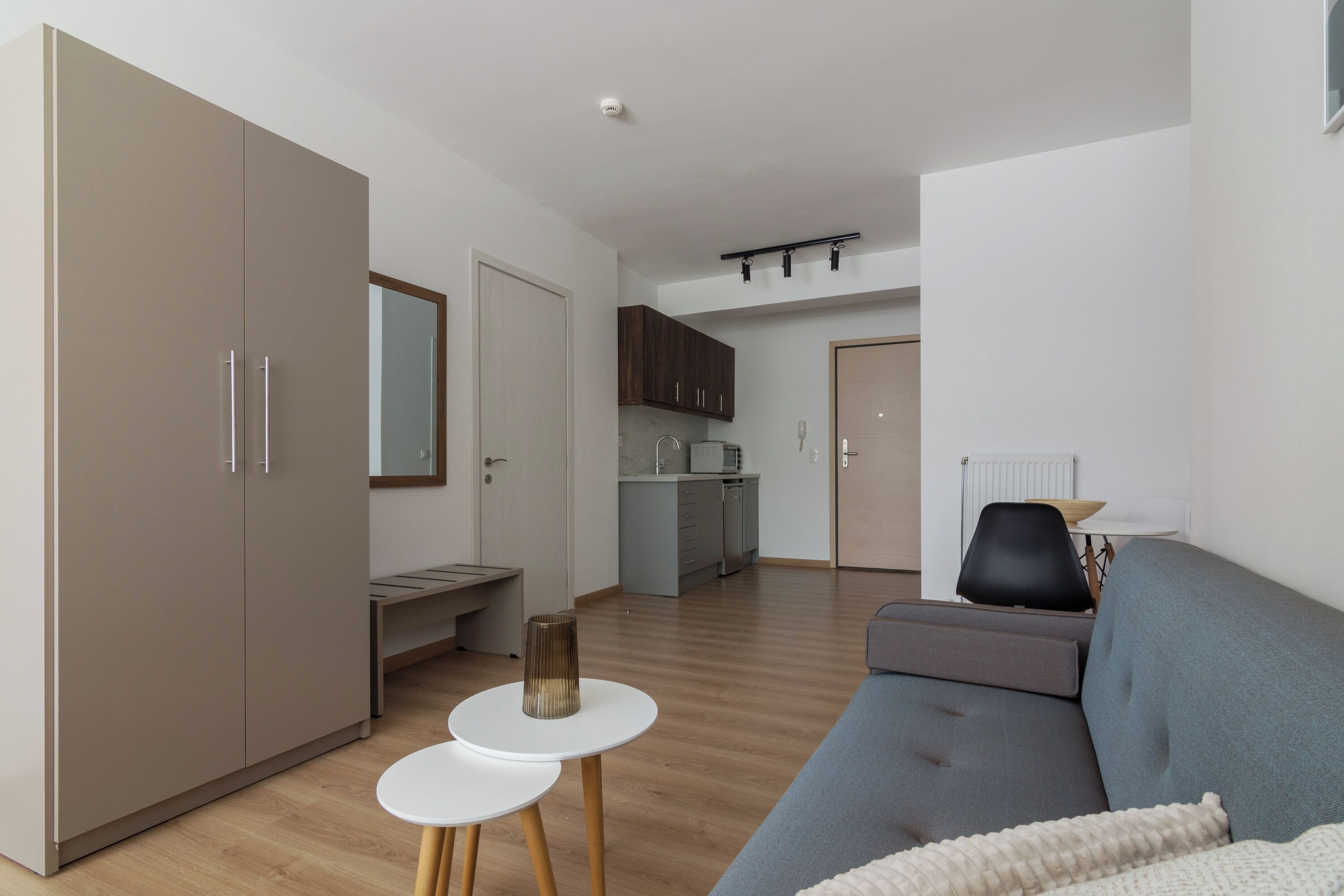 Ein modernes Wohnzimmer mit einem grauen Sofa, einem kleinen weißen Tisch und einem Kleiderschrank. Im Hintergrund ist eine Küche mit Schränken und einer Mikrowelle erkennbar.