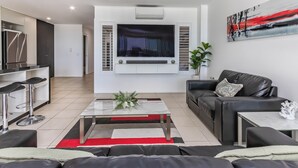 A modern living room with a flat-screen TV mounted on the wall, a black leather sofa, a glass coffee table, and a red and black striped rug.