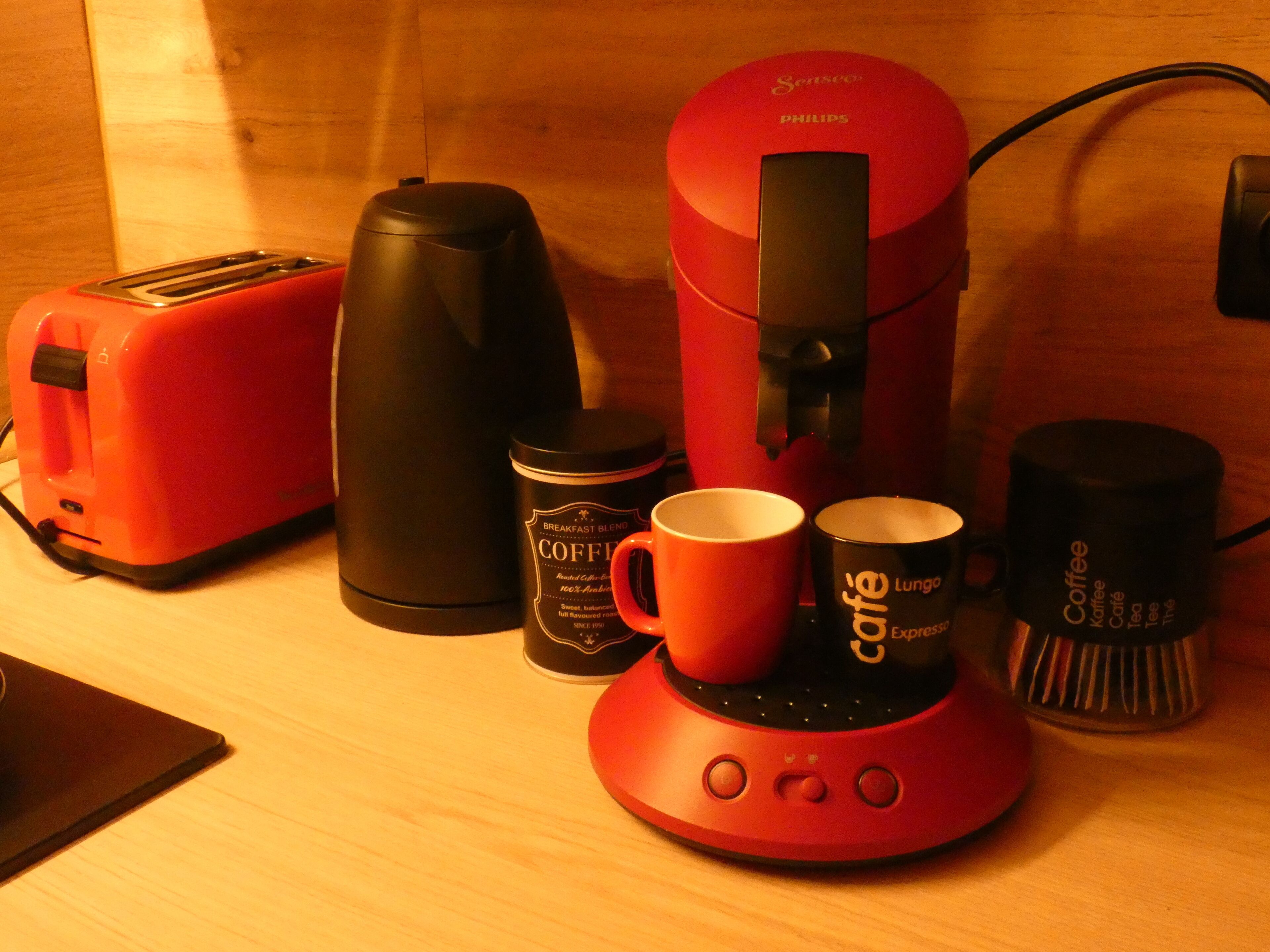 Un bancone cucina con un tostapane rosso, una macchina da caffè nera, una caffettiera rossa e due tazze da caffè.