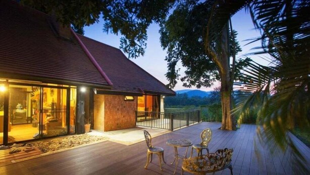 A wooden deck with outdoor furniture, a building with glass doors, and a view of the landscape. - Khaoyai Valley 4 (Pak Chong)