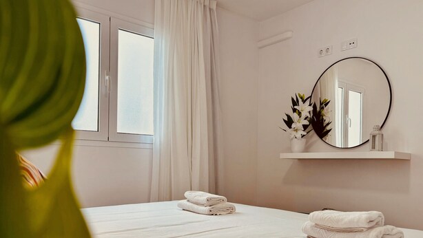Habitación de hotel con dos camas, una ventana con cortinas, un espejo redondo y una repisa con flores.