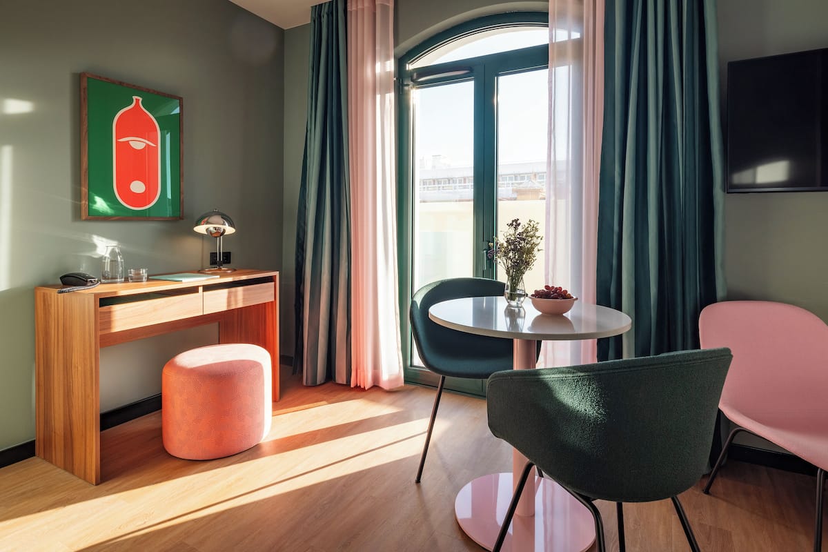 Een moderne hotelkamer met een houten bureau, een roze voetenbankje, een groene stoel, een klein tafeltje met een vaas en een schaal fruit, en een groot raam met gordijnen.