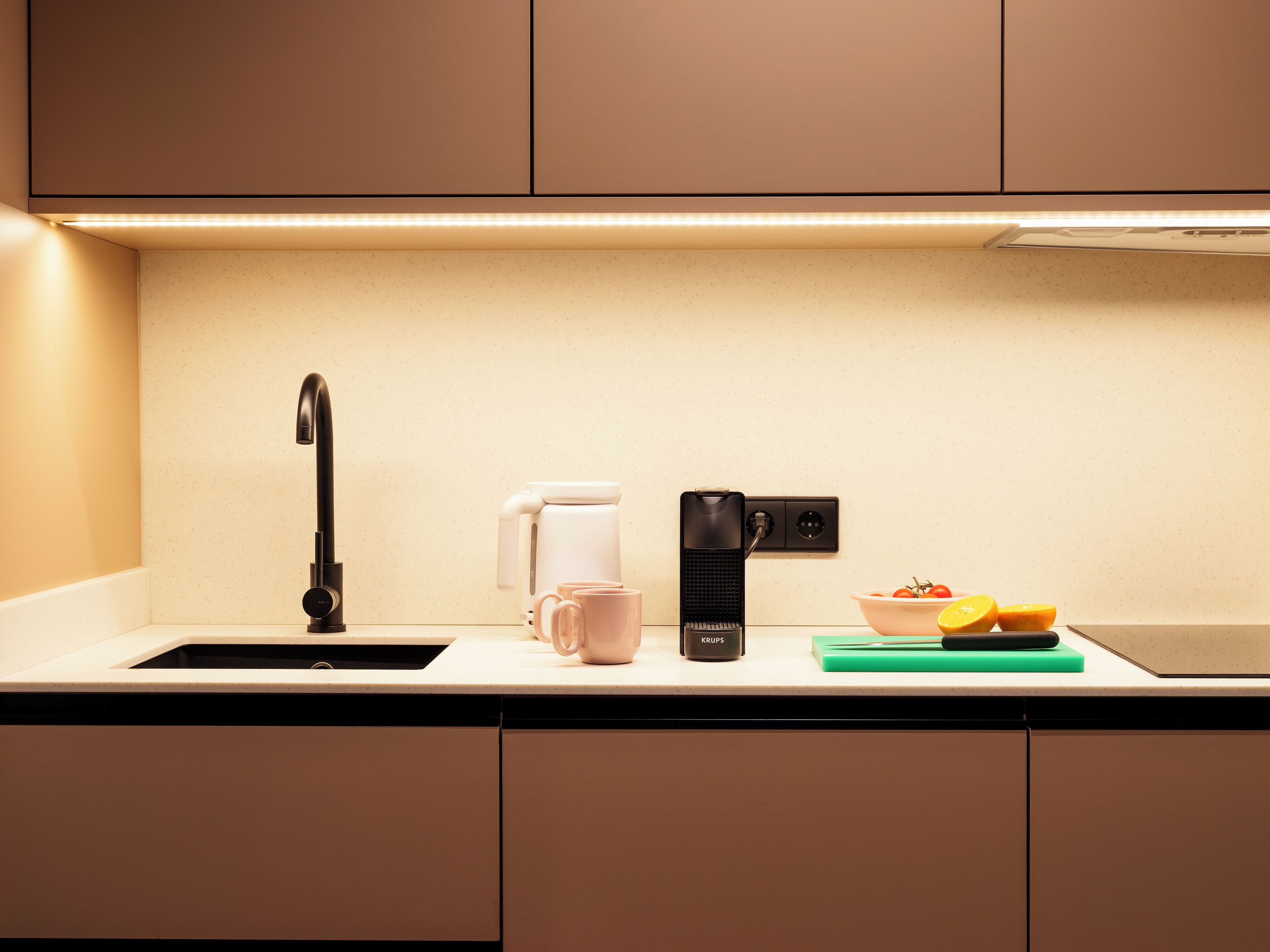 Een moderne keuken met een zwarte kraan, een witte koffiezetapparaat, een zwarte elektrische waterkoker en een schaal fruit op het aanrecht.