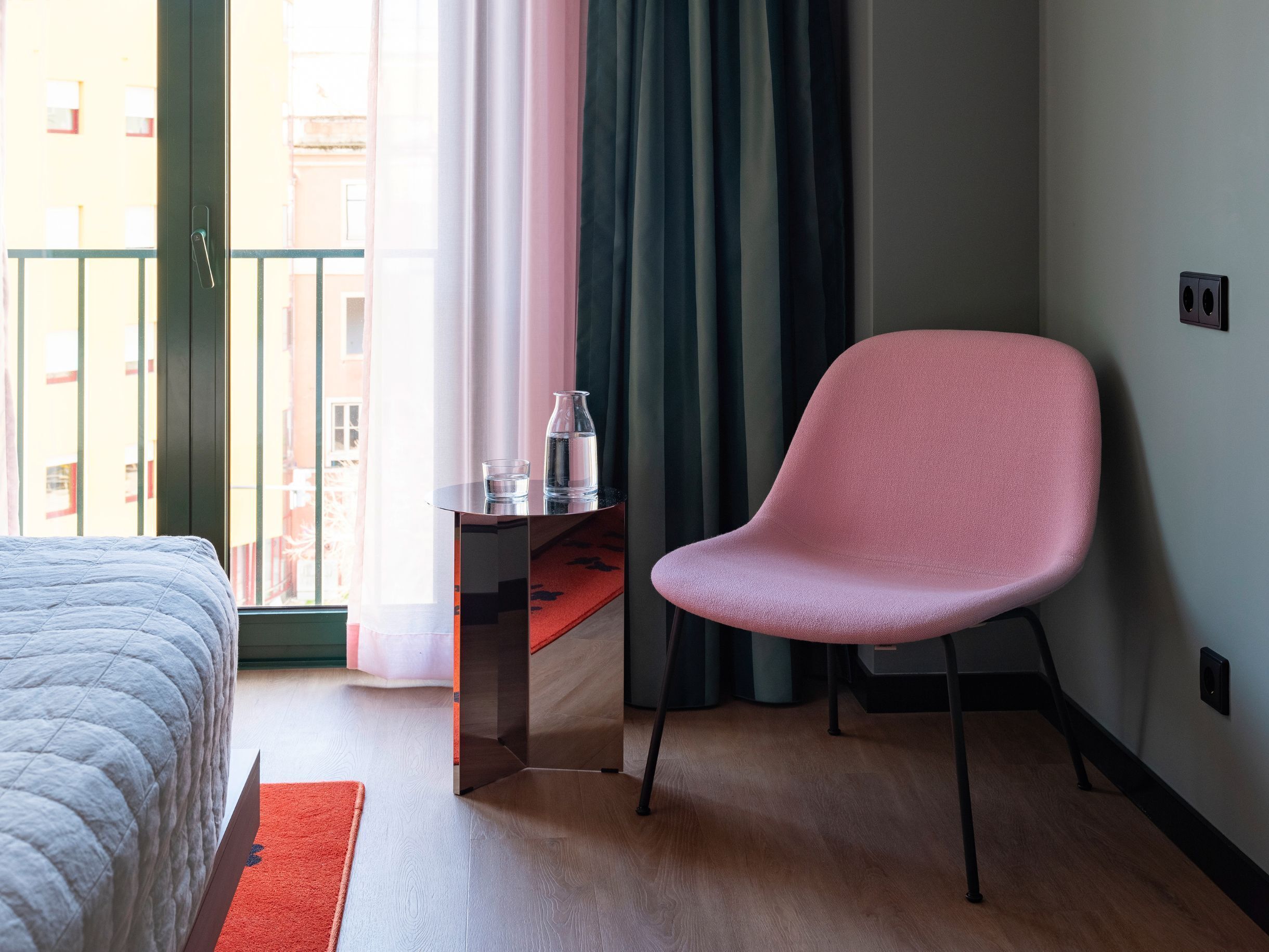 Habitación de hotel con cama, escritorio con silla, alfombra roja y vistas a edificios a través de la ventana.