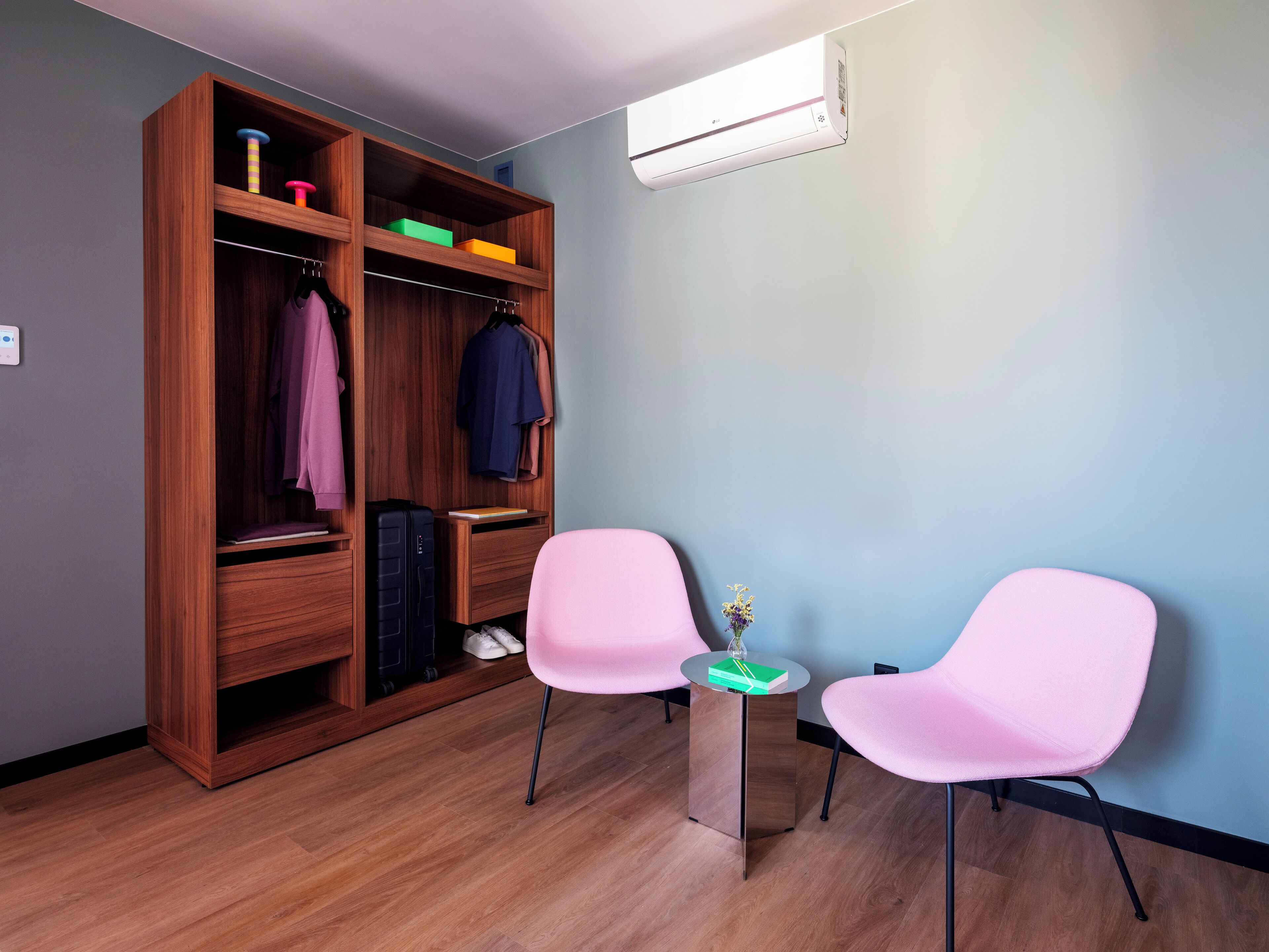 A room with a wooden wardrobe, two pink chairs, a small wooden table, and a wall-mounted air conditioner.