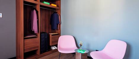 A room with a wooden wardrobe, two pink chairs, a small wooden table, and a wall-mounted air conditioner.