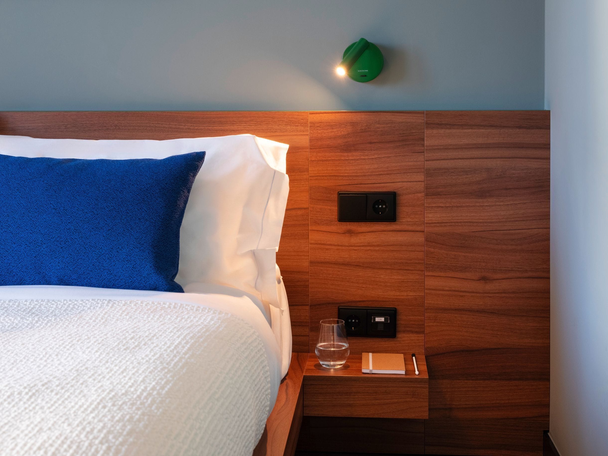 Een netjes opgemaakt bed met witte en blauwe beddengoed, een nachtkastje met een glas water, een notitieboekje en een pen, en een groen wandlamp.