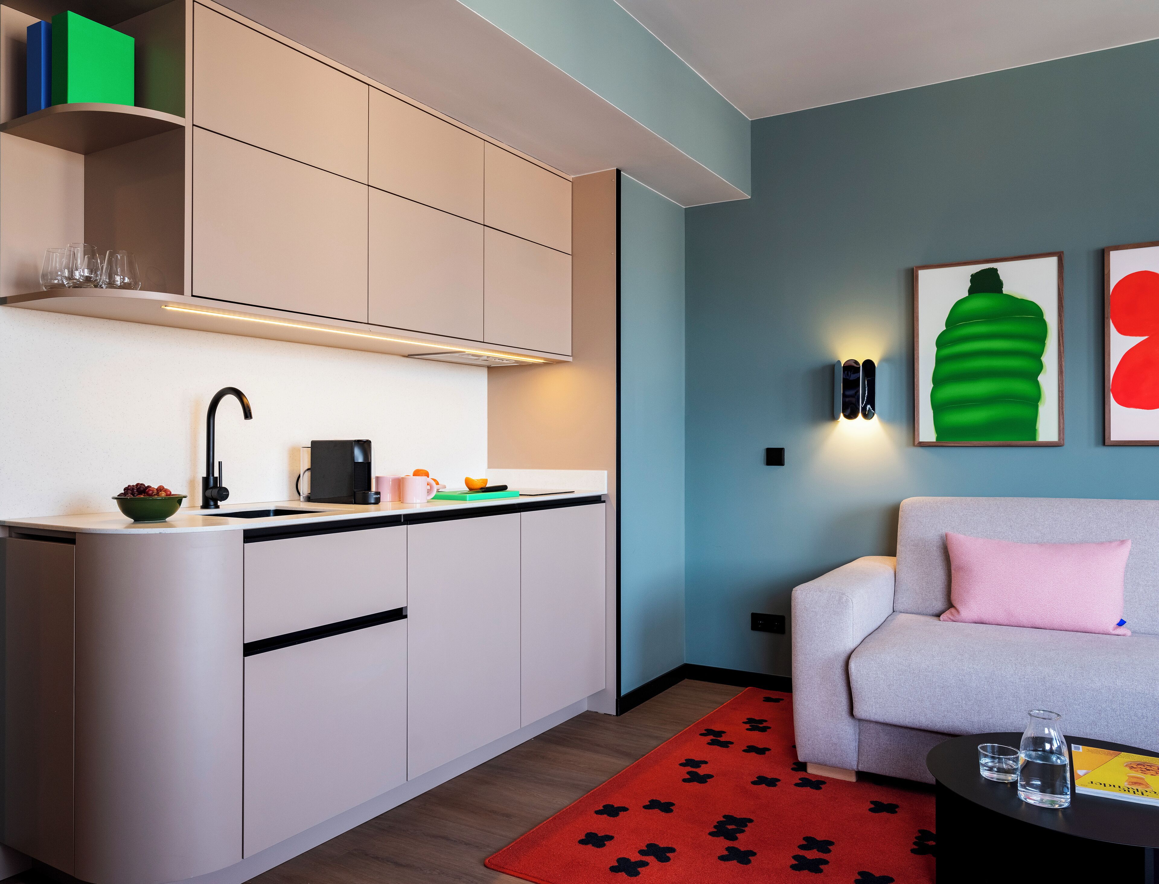 A modern kitchen with a built-in oven, a sink, and a countertop with a bowl of fruit and a coffee maker. There is a sofa with a pink pillow and a red rug in the living area.