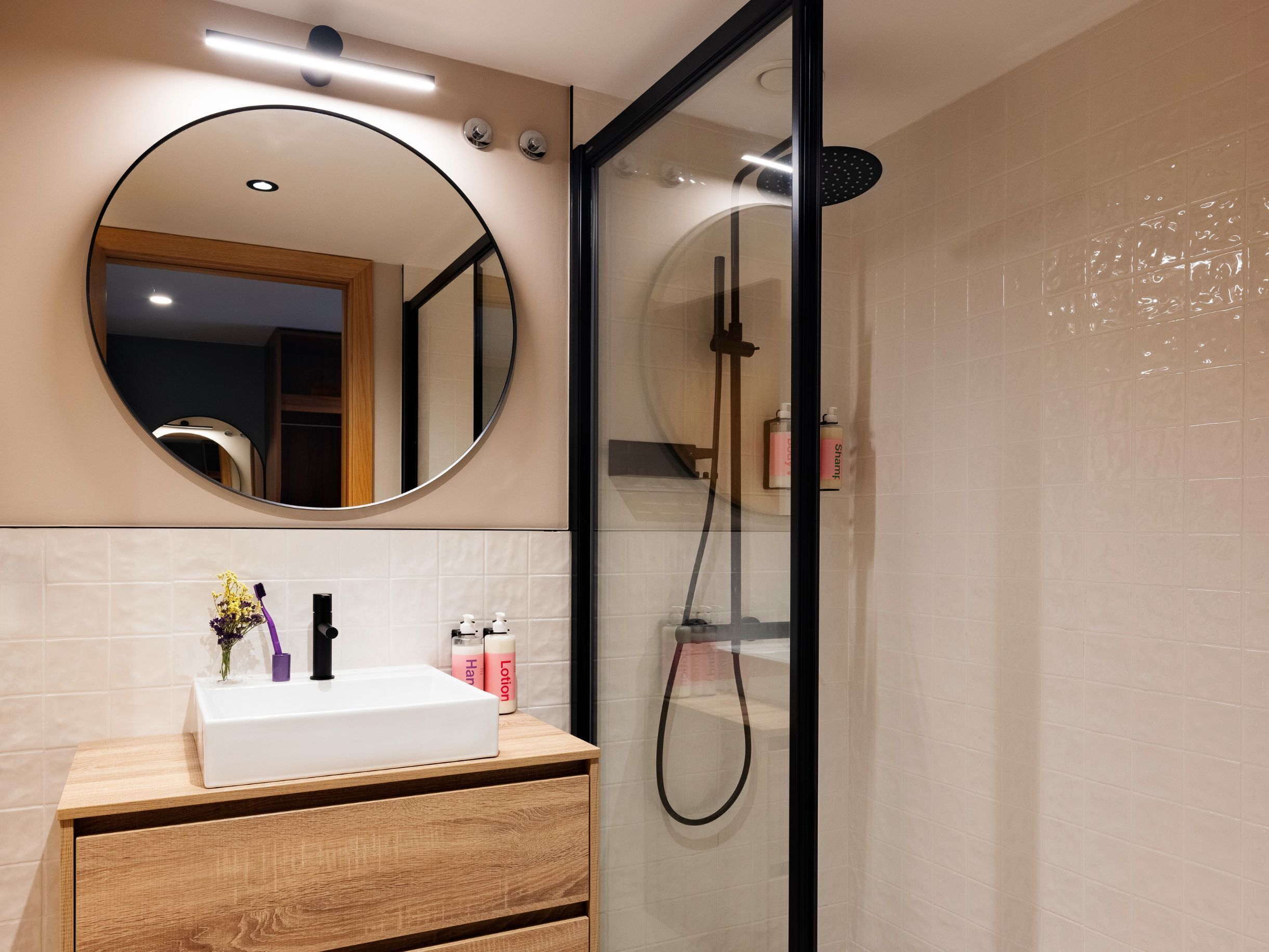 A modern bathroom with a glass-enclosed shower, a round mirror, and a wooden vanity with toiletries.