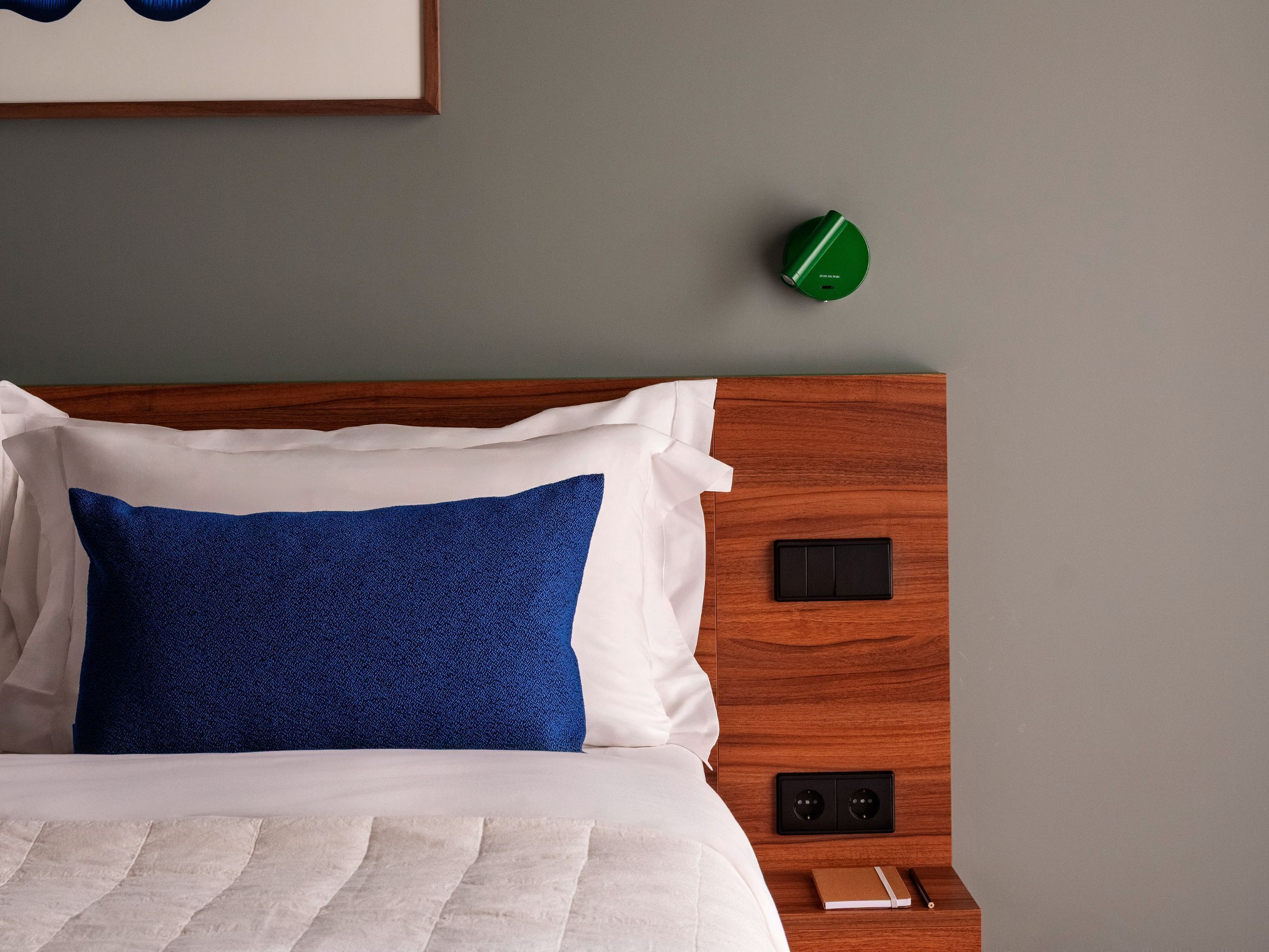 Un lit bien fait, avec des oreillers blancs et bleus, une tête de lit en bois, une table de chevet sur laquelle se trouvent un livre et un stylo, et une horloge verte accrochée au mur.