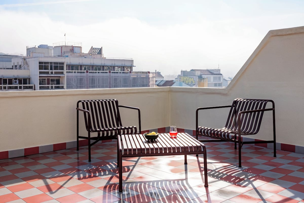 Een balkon met twee gestreepte stoelen en een kleine tafel, met een geruit vloerpatroon en een stadszicht op de achtergrond.
