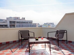 Ein Balkon mit zwei gestreiften Stühlen und einem kleinen Tisch, mit einem Schachbrettmuster auf dem Boden und Blick auf die Stadt im Hintergrund.