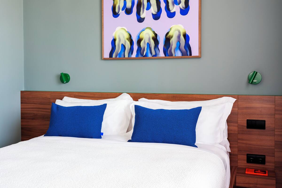 Een opgemaakt bed met wit- en blauw beddengoed, een houten hoofdbord en een aan de muur bevestigd schilderij.