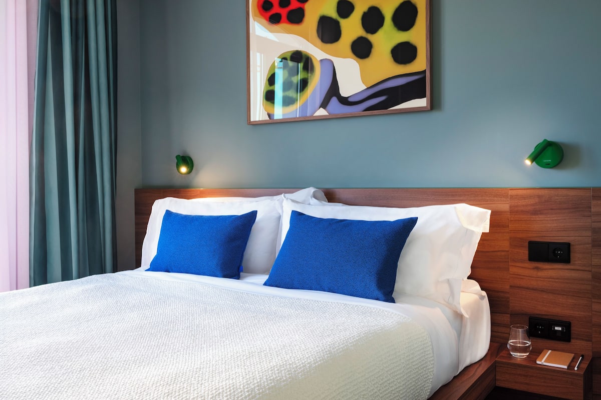Een net opgemaakt bed met wit- en blauwe beddengoed, een houten hoofdbord en een schilderij aan de muur.