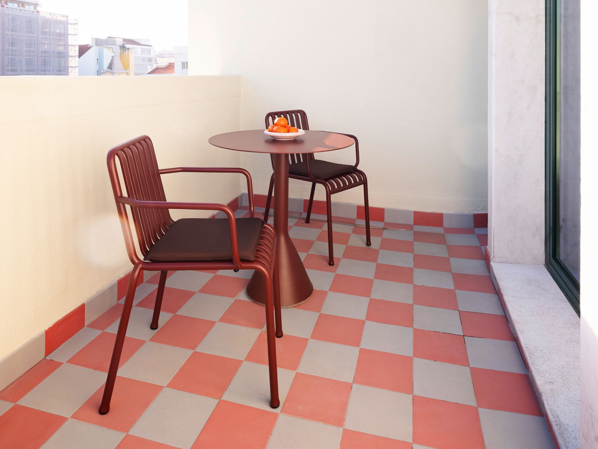 A small balcony with a checkered floor, two metal chairs, a small table, and a bowl of oranges.
