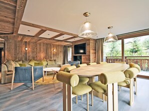 Un salon moderne avec un intérieur en bois, une table à manger ronde entourée de chaises, un téléviseur et une grande fenêtre donnant sur un paysage verdoyant.