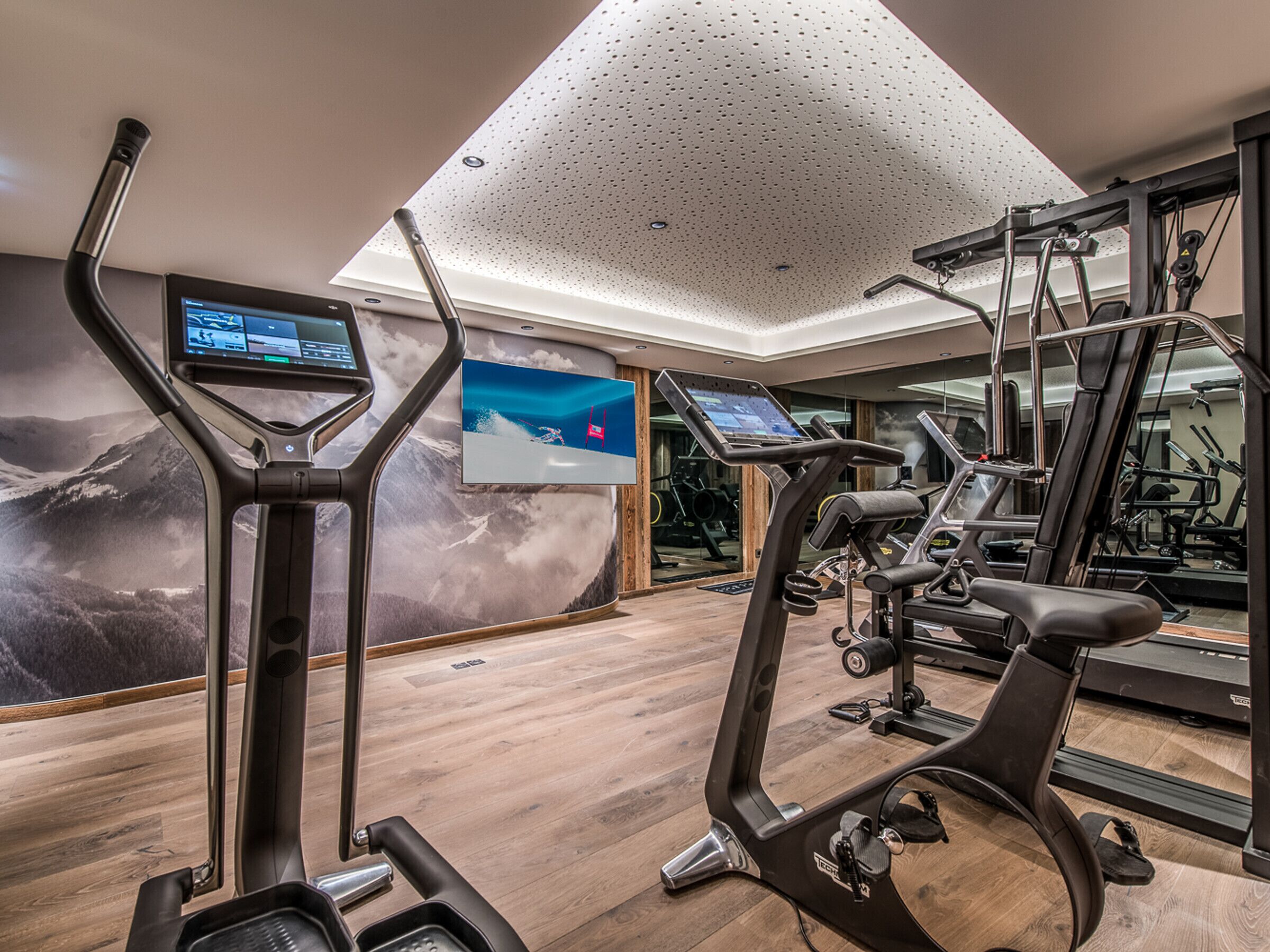 A modern gym with exercise equipment, a large screen, and a wooden floor.