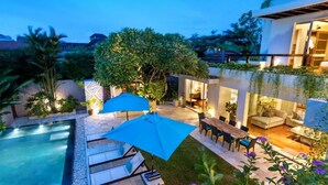 A modern outdoor pool area with lounge chairs and umbrellas, surrounded by lush greenery and a well-lit patio.