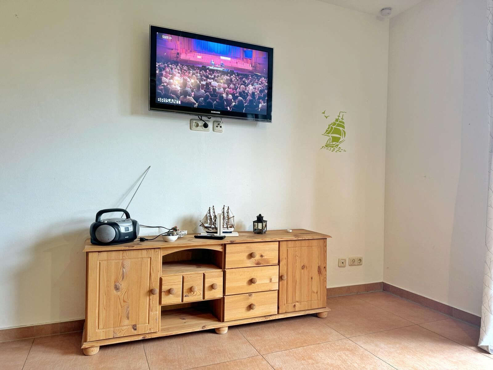 A wooden cabinet with drawers and a radio, a television mounted on the wall, and a small ship decoration on the wall.