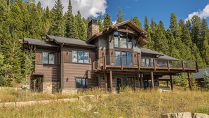 Une grande cabane en rondins à plusieurs étages, dotée d’une imposante cheminée en pierre, de grandes fenêtres et d’un balcon donnant sur une zone boisée.
